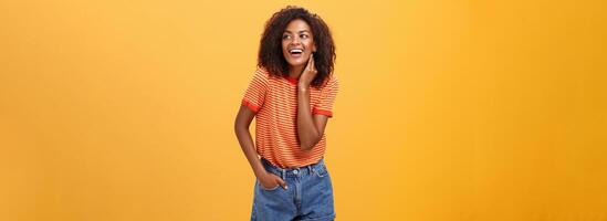 elegant vrouwelijk en modieus Afrikaanse Amerikaans vrouw model- met afro kapsel aanraken nek voorzichtig op zoek Rechtsaf met geamuseerd zorgeloos uitdrukking Holding hand- in zak- over- oranje achtergrond foto