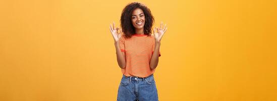 kilte aantrekkelijk tevreden Afrikaanse Amerikaans vrouw klant plukken nieuw kleding wezen verheugd en verheugd met koel personeel verhogen handen in Oke of geweldig gebaar glimlachen gelukkig over- oranje muur foto