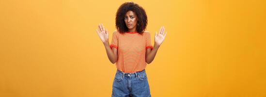 dwaas onzeker en verdrietig met een donkere huid vrouw model- in modieus gestreept t-shirt en shorts verhogen armen in overgave fronsen wezen niet betrokken en niet op de hoogte staand onwetend over- oranje achtergrond foto