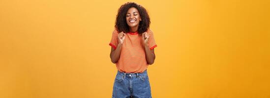 meisje gretig naar zien minnaar komt eraan van reis staand opgewonden en opgewonden met gelukkig breed glimlach balanceren vuisten grijnzend met Gesloten ogen gevoel blij en geamuseerd over- oranje achtergrond foto