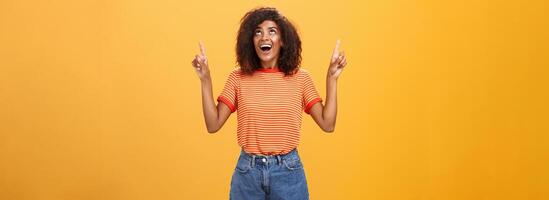 gefascineerd onder de indruk en geamuseerd mooi charmant Afro-Amerikaans vrouw met afro kapsel in modieus t-shirt en denim shorts richten en op zoek omhoog met geïnteresseerd kijken over- oranje muur foto