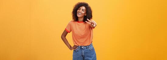 vriendelijk zorgeloos en gelukkig mooi hoog Afrikaanse Amerikaans vrouw model- met gekruld kapsel Holding hand- Aan heup kantelen hoofd en glimlachen blij tonen vrede gebaar over- oranje muur foto