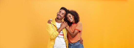 ze liefde elk ander. portret van twee charmant Afrikaanse Amerikaans Mens en vrouw in verhouding knuffelen met hartverwarmend glimlach aanraken handen glimlachen voorzichtig poseren tegen oranje achtergrond foto