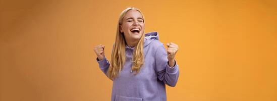 schattig gelukkig zegevieren blond Europese meisje in capuchon schreeuwen Ja bereiken droom levenslang klemmen vuisten gelukkig dansen vieren zege succes bereiken doel, staand oranje achtergrond foto