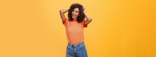 rebels speels en blij Afrikaanse Amerikaans meisje met gekruld kapsel maken duivel hoorns Aan hoofd met inhoudsopgave vingers glimlachen breed tonen waaghals karakter en charisma over- oranje muur foto