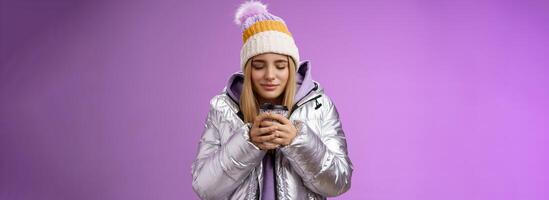 charmant inschrijving romantisch blond meisje snuiven mooi hoor geur warm smakelijk koffie Holding afhalen kop warm handen dichtbij ogen kijken verheugd vervelend winter jasje hoed genieten vakantie, Purper achtergrond foto