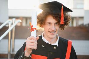 diploma uitreiking van Universiteit. jong glimlachen jongen Universiteit afstuderen in traditioneel botten en mantel staand en Holding diploma in hand- over- Universiteit gebouw achtergrond foto