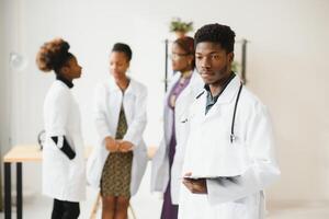 jong Afrikaanse mannetje dokter glimlachen terwijl staand in een ziekenhuis gang met een verschillend groep van personeel in de achtergrond. foto