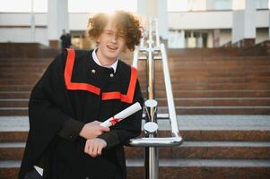 diploma uitreiking van Universiteit. jong glimlachen jongen Universiteit afstuderen in traditioneel botten en mantel staand en Holding diploma in hand- over- Universiteit gebouw achtergrond foto