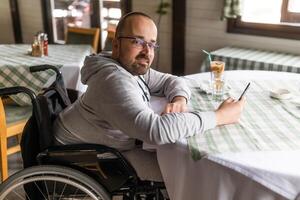 gelukkig verlamd gehandicapten Mens in rolstoel is zittend Bij restaurant en gebruik makend van smartphone. foto
