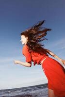 strand vreugde. een glimlachen vrouw dansen met vreugde in rood kleren, genieten van de vrijheid van zomer door foto