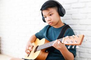 Aziatisch jongen vervelend hoofdtelefoons met zwart muziek- zitten en Speel gitaar in de huis. muziek- aan het leren concept, muziek- vaardigheid opleiding. kopiëren ruimte foto