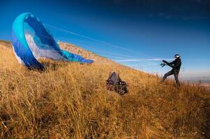 de paraglider is voorbereidingen treffen naar nemen uit. een kleurrijk vlieger leugens Aan de gras, in de achtergrond Daar is een mooi landschap, Doorzichtig blauw lucht foto
