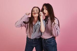 snor maken met haar. jonge vrouwen die plezier hebben in de studio met roze achtergrond. schattige tweeling foto