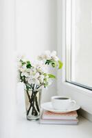 wit kop van koffie Aan een wit bord, een stack van boeken en een vaas van bloemen Aan de vensterbank knus Pasen, voorjaar nog steeds leven. foto