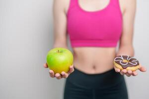 vrouw hand- houden groen appel en donut, vrouw geschiktheid Kiezen tussen fruit is gezond en zoet is ongezond rommel voedsel. dieet controle, gewicht verlies, zwaarlijvigheid, aan het eten levensstijl en voeding concept foto