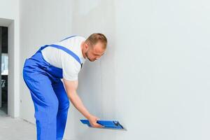 de bouwer voorzichtig corrigeert de onregelmatigheden van de muur met een troffel. bouwer in werk kleren tegen een grijs muur. foto stukadoor Bij werk.