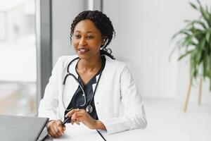 geneesmiddel, online onderhoud en gezondheidszorg concept - gelukkig glimlachen Afrikaanse Amerikaans vrouw dokter of verpleegster met koptelefoon en laptop hebben conferentie of video telefoontje Bij ziekenhuis. foto