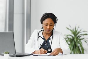 geneesmiddel, online onderhoud en gezondheidszorg concept - gelukkig glimlachen Afrikaanse Amerikaans vrouw dokter of verpleegster met koptelefoon en laptop hebben conferentie of video telefoontje Bij ziekenhuis. foto