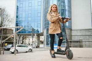 mooi jong vrouw in zonnebril is zittend Aan haar elektro scooter in de buurt groot glas gebouw. foto