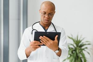 volwassen Afrikaanse dokter gebruik makend van digitaal tablet in gang . portret van zelfverzekerd mannetje dokter gebruik makend van tablet computer in kliniek met kopiëren ruimte. geslaagd glimlachen dokter in Lab jas vervelend stethoscoop foto