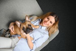 jong mooi vrouw in gewoontjes kleren knuffelen haar hond zittend Aan de sofa in de leven kamer van haar knus land huis. dier communicatie concept foto