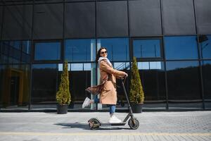 jong vrouw Aan de elektrisch scooter Aan de achtergrond van kantoor gebouw, scooter foto