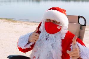 de kerstman claus verwijdert een rood medisch masker aan het liegen Aan een zon ligstoel Aan de strand. einde van de epidemie foto