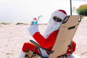 de kerstman claus in zonnebril drankjes een cocktail terwijl aan het liegen Aan een zon ligstoel Aan de meer strand. de kerstman in de tropen foto