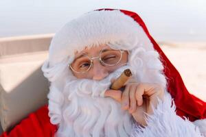 de kerstman in bril rookt een sigaar terwijl aan het liegen Aan een zon ligstoel Aan een meer strand foto