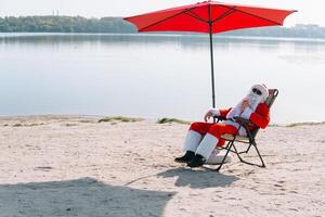 de kerstman claus in zonnebril drankjes een cocktail terwijl aan het liegen Aan een zon ligstoel Aan de meer strand. de kerstman in de tropen foto