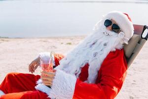 de kerstman claus in zonnebril drankjes een cocktail terwijl aan het liegen Aan een zon ligstoel Aan de meer strand. de kerstman in de tropen foto