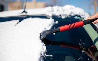 een jong Mens vervelend zwart warm pak voor reinigt zijn auto na een sneeuwval Aan een zonnig, ijzig dag. schoonmaak en opruimen de auto van sneeuw Aan een winter dag en een erge, ernstige sneeuwstorm in winter. foto