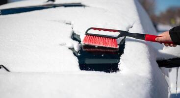 een jong Mens vervelend zwart warm pak voor reinigt zijn auto na een sneeuwval Aan een zonnig, ijzig dag. schoonmaak en opruimen de auto van sneeuw Aan een winter dag en een erge, ernstige sneeuwstorm in winter. foto