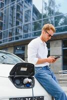 Mens Holding macht opladen kabel voor elektrisch auto in buitenshuis auto park. en hij s gaan naar aansluiten de auto naar de opladen station in de parkeren veel in de buurt de boodschappen doen centrum foto