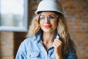 aantrekkelijk vrouw bouw arbeider in harde hoed. zelfverzekerd jong specialist in geruit blauw overhemd in jeans staand in leeg kamer. interieur ontwerp en vernieuwing onderhoud. foto