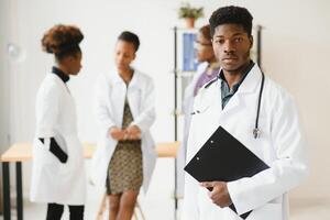 jong Afrikaanse mannetje dokter glimlachen terwijl staand in een ziekenhuis gang met een verschillend groep van personeel in de achtergrond. foto