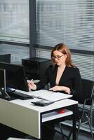 mooi, leuk, schattig, perfect vrouw zittend Bij haar bureau Aan leer stoel in werk station, vervelend bril, formele kleding, hebben laptop en notitieboekje Aan de tafel foto