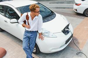 Mens Holding macht opladen kabel voor elektrisch auto in buitenshuis auto park. en hij s gaan naar aansluiten de auto naar de opladen station in de parkeren veel in de buurt de boodschappen doen centrum foto