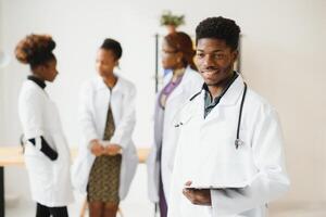 jong Afrikaanse mannetje dokter glimlachen terwijl staand in een ziekenhuis gang met een verschillend groep van personeel in de achtergrond. foto