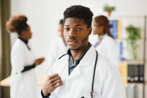 jong Afrikaanse mannetje dokter glimlachen terwijl staand in een ziekenhuis gang met een verschillend groep van personeel in de achtergrond. foto