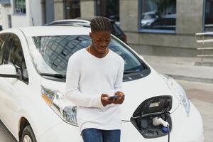 Afrikaanse Mens gebruik slim telefoon terwijl aan het wachten en macht levering aansluiten naar elektrisch voertuigen voor opladen de accu in auto foto
