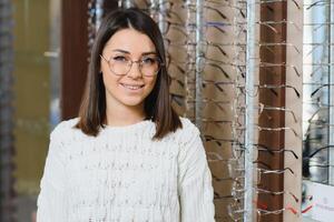 vrouw kiest bril in de op te slaan. brunette in een wit trui koopt bril. meisje Aan een achtergrond van winkel ramen met verschillend modellen van bril foto