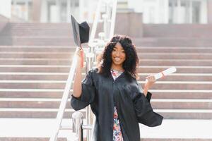 vrolijk afro Amerikaans vrouw afstuderen staand in voorkant van Universiteit gebouw foto