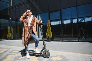 jong mooi vrouw in een jasje glimlacht en ritten een elektrisch scooter naar werk langs kantoor gebouwen foto