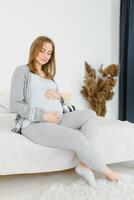 zwanger jong Europese vrouw zittend Aan een groot bed en strelen haar groot buik, 7 maand zwangerschap, aan het wachten voor de geboorte van haar baby. foto