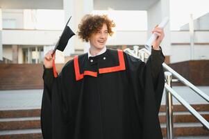 gelukkig glimlachen Universiteit afstuderen in mantel Holding diploma in verheven hand- en uitdrukken geluk over- Universiteit gebouw Bij achtergrond. geslaagd afstuderen van Universiteit of college foto