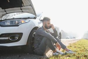 Mens gebruik een mobiele telefoon telefoontje garage in voorkant van de Open kap van een gebroken auto Aan de weg in de Woud. auto afbreken concept. foto