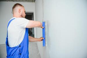 portret van een arbeider in overall en Holding een stopverf mes in zijn handen tegen de gepleisterd muur achtergrond. reparatie werk en bouw concept foto