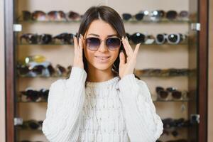 welke een is beter fit mij. portret van vrolijk aarzelend vrouw in opticien op te slaan, maken beslissing, Holding elegant zonnebril, kiezen wat ze zou moeten kopen. Super goed kortingen voor buying twee paren. foto